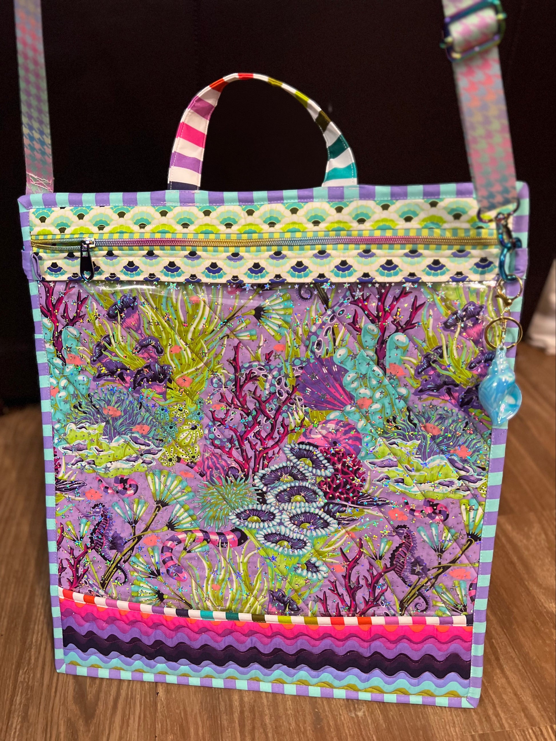 A square tote bag with a colorful, busy tropical fish and coral print, featuring a patterned top border, a zippered top closure, and a striped shoulder strap, shown on a wooden floor.